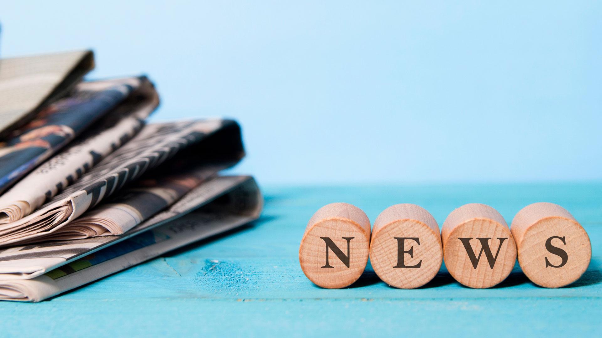 NEWS word in small wooden circles, with a pile of newspapers next to it, on a blue background.