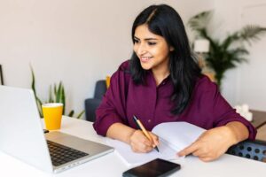 Young adult Indian woman taking notes while using laptop