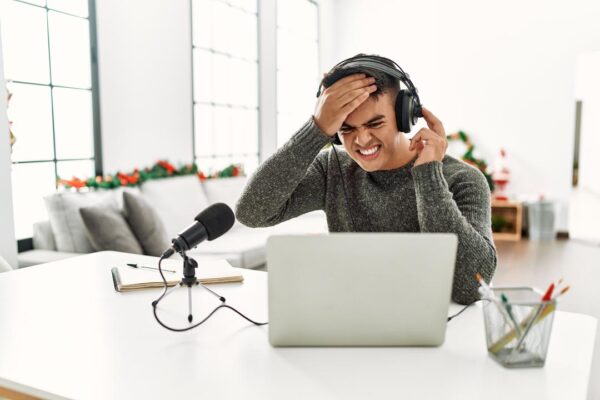 Hispanic man recording podcast stressed and frustrated with hand on head.