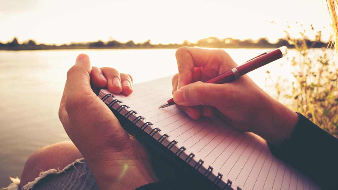 Close up hand of young woman with pen writing on notebook at a river.