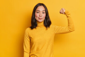 Serious self confident brunette young Asian woman raises hand up and shows demonstrates strength looks like strong powerful bodybuilder dressed in casual turtleneck isolated over yellow background
