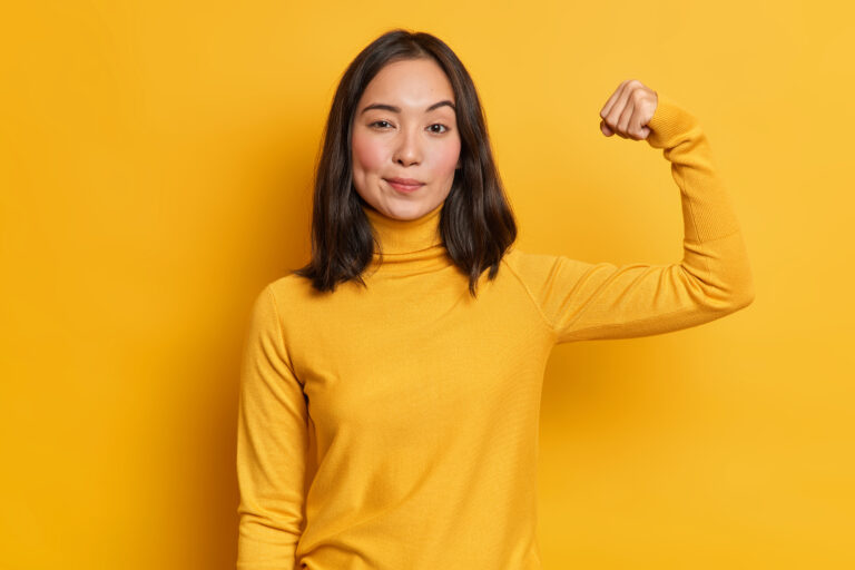 Serious self confident brunette young Asian woman raises hand up and shows demonstrates strength looks like strong powerful bodybuilder dressed in casual turtleneck isolated over yellow background