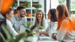 A group of five business professionals are sitting around a table having a meeting. They are all smiling and appear to be engaged in the conversation. There is a potted plant on the table. The