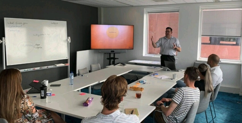 Andy McConville standing in a blue shirt and suit trousers, delivering training to a small group of people, with a TV monitor showing a slide and a whiteboard with writing on it. (C) Andy McConville