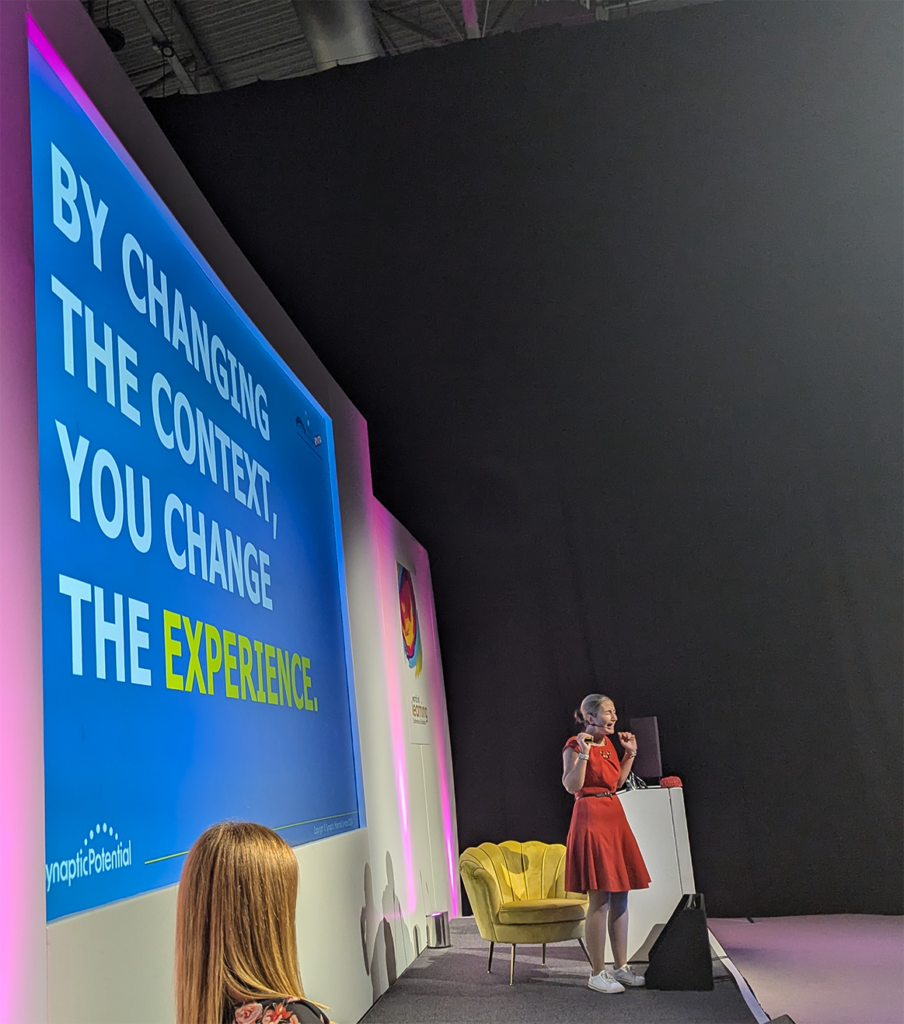 Slide at a conference reading "By changing the context, you change the experience", with Amy Brann on stage looking excited