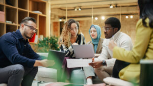A Group of Multiethnic People Brainstorming Using Laptop and Charts in a Meeting Room at Office. Enthusiastic Young Adults Analyzing New Data To Improve Startup Performance. Medium Shot