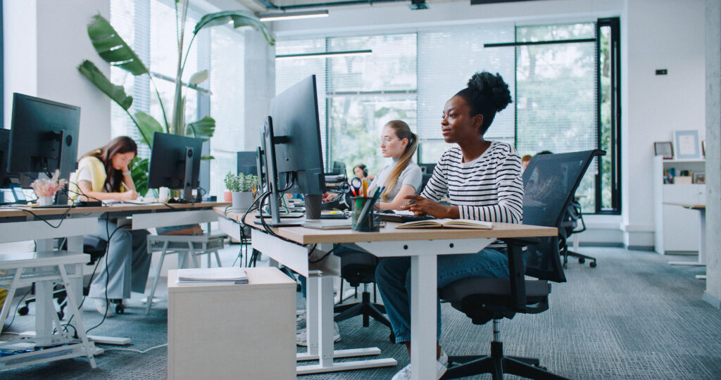 Zooming in on pretty woman with casual clothes working at computer in office. Distracted by camera from work and joyfully smiling. In background people sitting at their workplaces. Job concept.