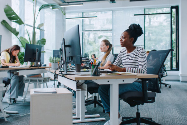 Zooming in on pretty woman with casual clothes working at computer in office. Distracted by camera from work and joyfully smiling. In background people sitting at their workplaces. Job concept.