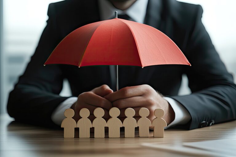 Businessman protecting team under red umbrella.  Hands hold umbrella over small wooden figures of people on a desk