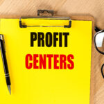 Clipboard with text profit centers on bright yellow paper next to pen and eyeglasses on wooden table