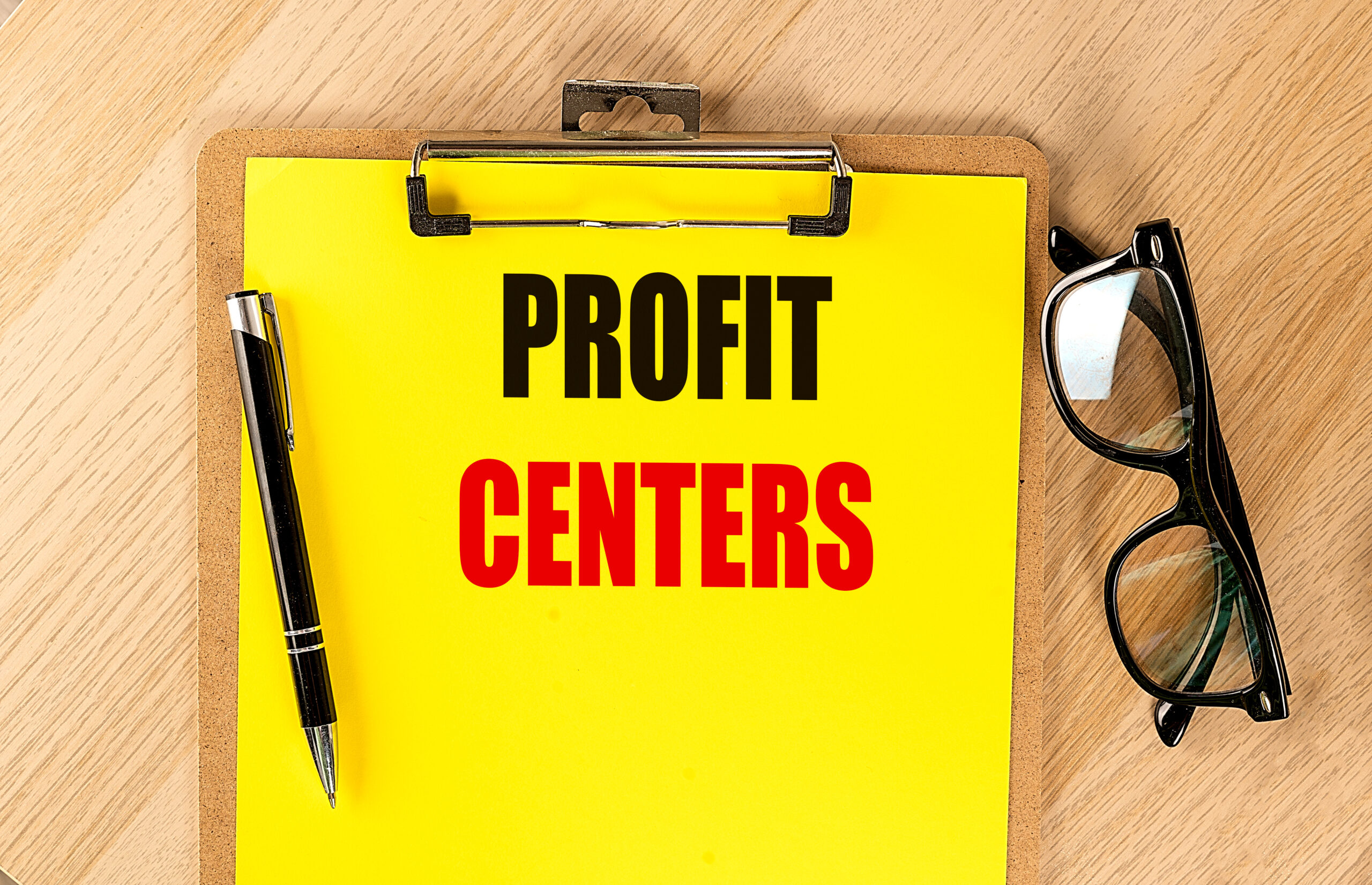Clipboard with text profit centers on bright yellow paper next to pen and eyeglasses on wooden table