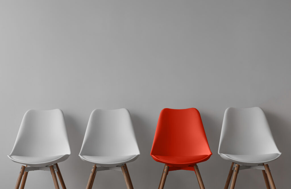 Three white chairs and red on gray wall background in office or room. Row of stools with one of different colour. Job opportunity, recruitment, business, leadership and hiring concept, copy space