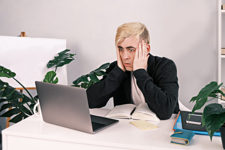 A young, frustrated, shocked man holds on to his head staring at a laptop screen with his eyes wide open while sitting at his desk in the office. Bad news, , failure in work.
