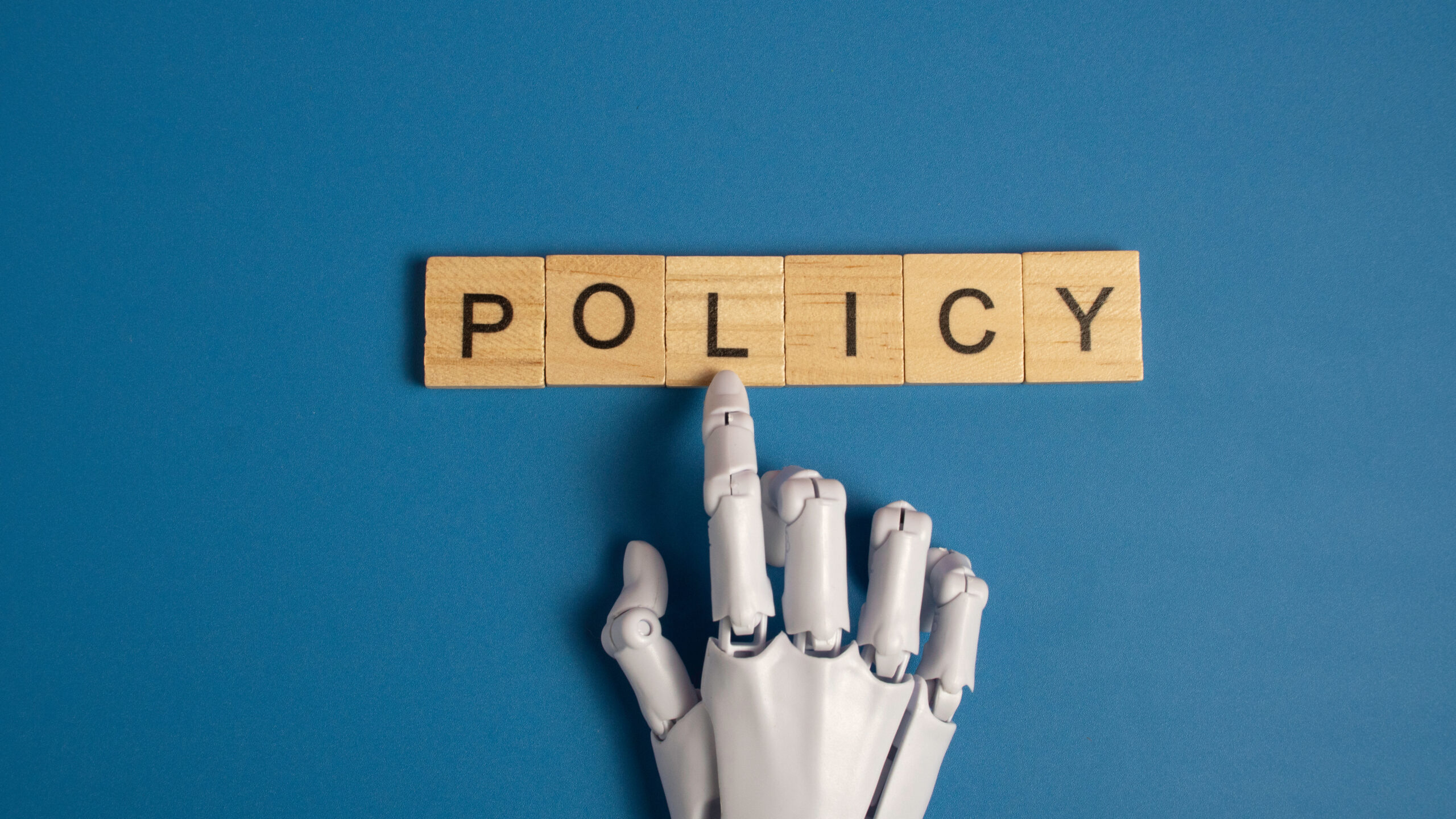 White robotic hand pointing to the word policy on wooden blocks over a blue background representing artificial intelligence ethics and government regulations.
