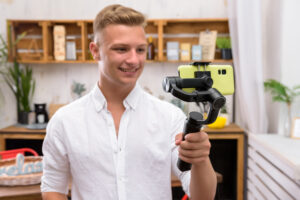 Young handsome guy filming a video blog on a mobile device using an electronic image stabilizer in the kitchen. Blond European man shoots a video in a good mood