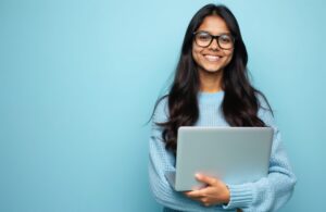 Young Indian woman with glasses smiles holding laptop. She wears blue sweater. Confident student ready for work or study. Person works on computer against blue background.