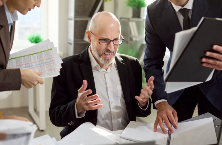 Frustrated manager with colleagues under deadline pressure. Paperwork and overload spark stress and frustration as the team crowds his desk, fueling burnout. Office stress and burnout concept.