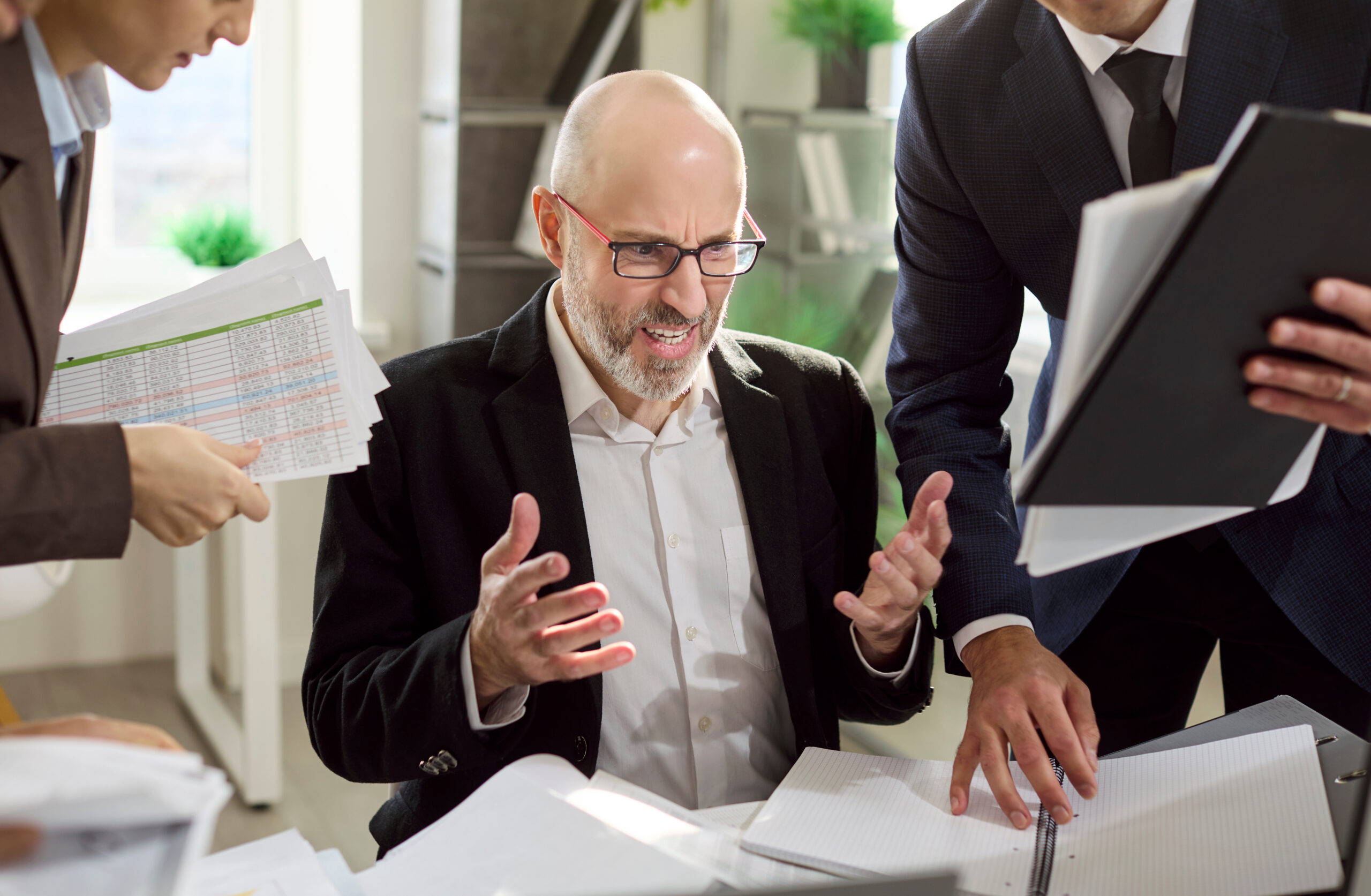 Frustrated manager with colleagues under deadline pressure. Paperwork and overload spark stress and frustration as the team crowds his desk, fueling burnout. Office stress and burnout concept.