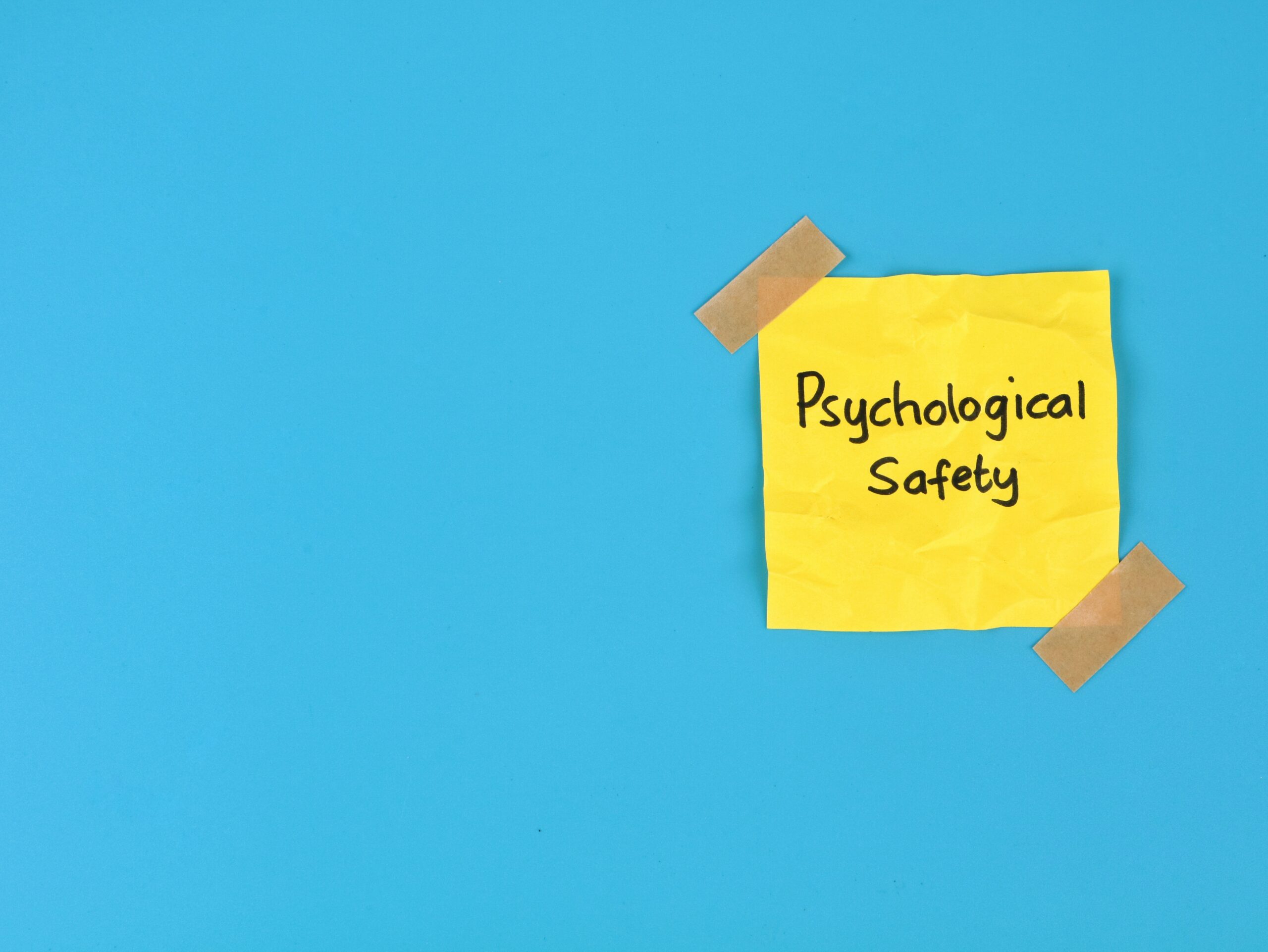 Psychological safety text handwritten on yellow adhesive note over blue background. Perfect for business management, positive workplace culture, mental health, teamwork, and employee well-being